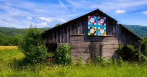 A Brown Old-School Barn Set in a Green and Yellow Field Against a Backdrop of Rising Green Mountains, has a very Multi Colored Image on its Roof.